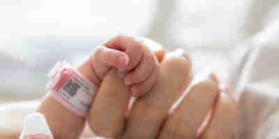 A newborn baby's arm and hand with identification band on its wrist, holding an adult hand. Continuing the conversation around birth trauma and childbirth-related PTSD