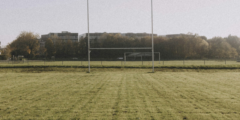 An image of a rugby pitch in a park. Rugby goal posts are directly in front and approximately 30 meters away. There are tress in the background and four flats flat roofs visible beyond.