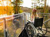 Young girl in a wheelchair on a balcony overlooking a river and tall trees. Spinal cord injury compensation claim solicitors